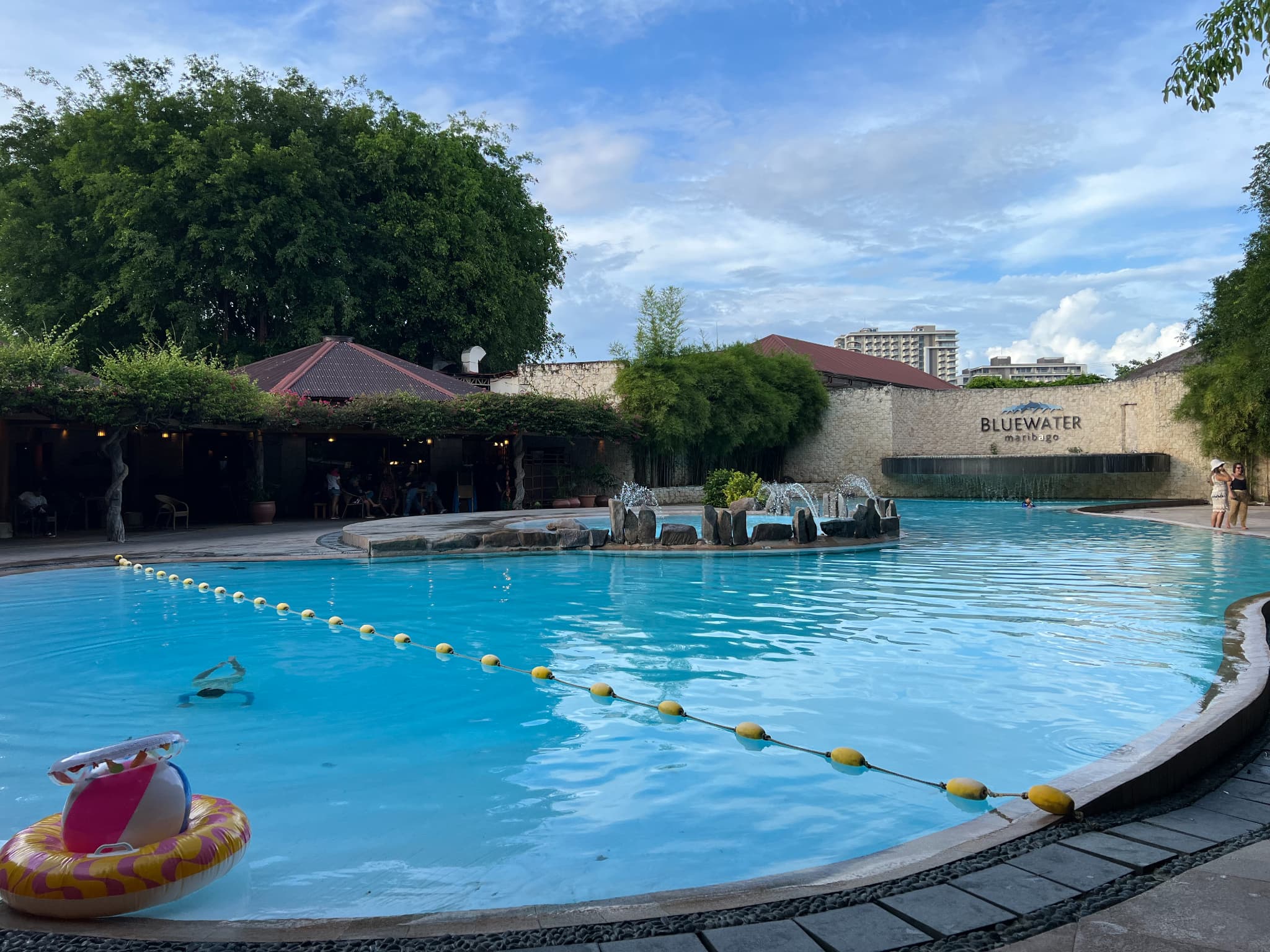 ブルーウォーターマリバゴのメインプールの全景。手前には浮き輪が浮かび、奥には石造りのウォーターフェティチャー（噴水）と、ホテルのロゴ『BLUEWATER maribago』が刻まれた白い壁が見える。夕暮れ時の青空と大きな木々がリゾート感を演出している。