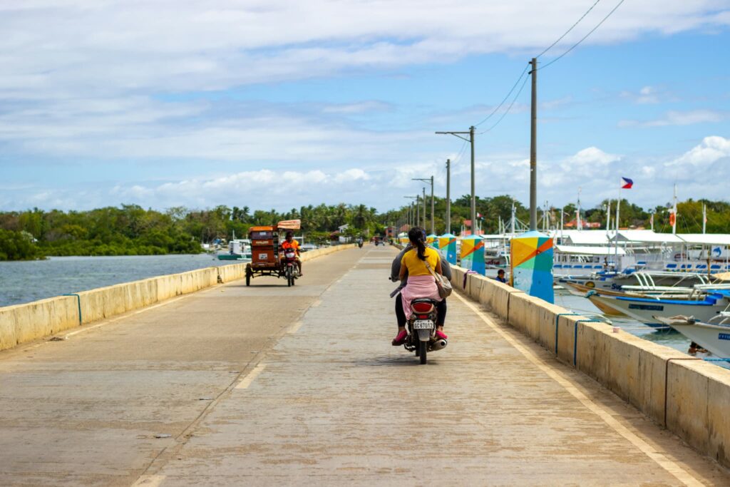 晴れた日のフィリピン、セブ島のコンクリート製の桟橋を走るオートバイの二人乗り。桟橋の右側には青い海が広がり、多数のボートが停泊している。左側には緑豊かなマングローブ林が見える。