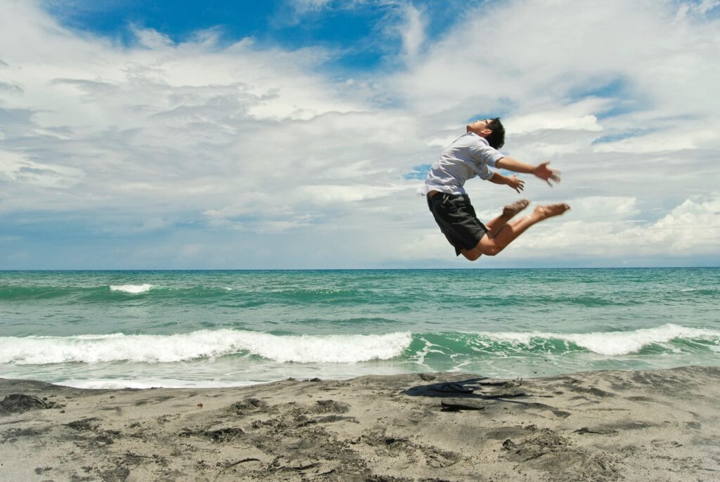 青い海と空を背景に、砂浜で大きくジャンプをして喜びを表現している男性。